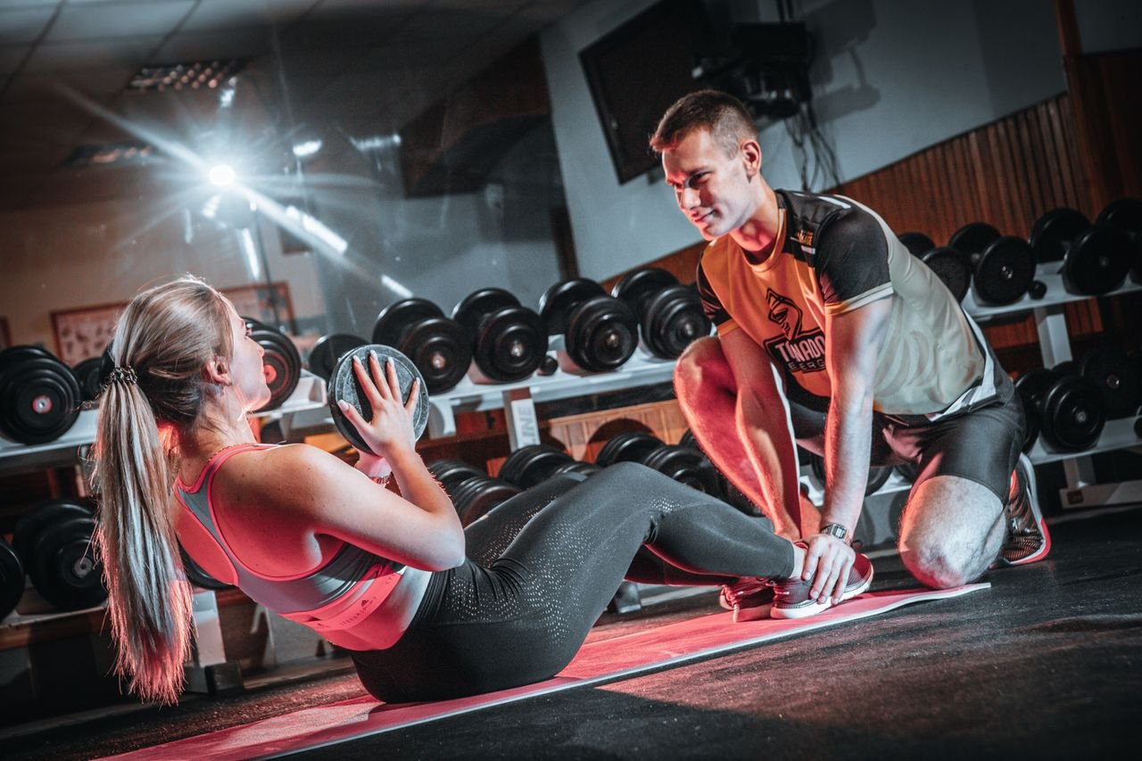 The Art of Drawing Readers In: Your attractive post title goes here A personal trainer assists a woman doing sit-ups with a medicine ball in a gym setting.